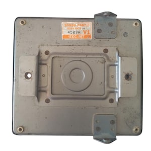 May include: A rectangular, silver-coloured automotive computer module with mounting brackets. The module has a central circular indentation and a sticker with text including "4509A V1" and "EEC-MT".