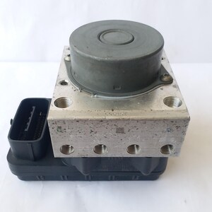 Op de afbeelding: Een zilverkleurig auto-onderdeel met een cilindrische bovenkant en meerdere schroefdraadaansluitingen. Een zwart rechthoekig onderdeel met een connector is aan de basis bevestigd. Het onderdeel lijkt van metaal en kunststof te zijn.