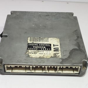 May include: A silver Toyota engine control module with a white label bearing black text. The label states "ENGINE CONTROL TOYOTA 89661-26861 75800-3650 12V DENSO MADE IN JAPAN". The module features a white connector.