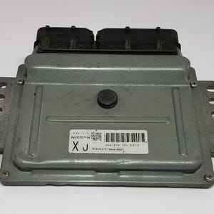 May include: A silver Nissan engine control module (ECM) with black connectors. The ECM has a label with text and a barcode. The unit has a rectangular shape with mounting tabs and a central button.