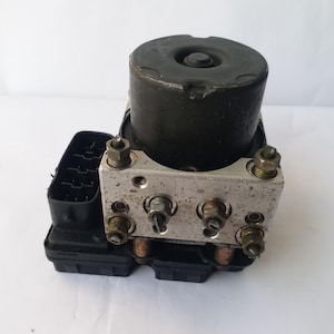 Op de afbeelding: Een ABS-module (antiblokkeersysteem) voor auto's. De unit bestaat uit een zwart cilindrisch component aan de bovenkant, een zilverkleurig metalen blok met diverse fittingen en een zwarte kunststof behuizing met elektrische connectoren.