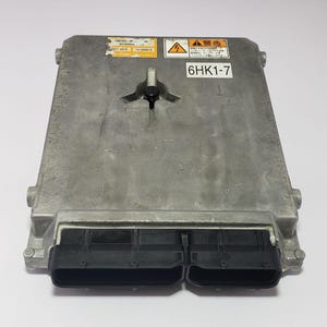 Puede incluir: Una unidad de control electrónico (ECU) de color plateado con conectores negros. La ECU tiene varias pegatinas con texto y el número "6HK1-7" impreso. La unidad es rectangular con esquinas redondeadas.