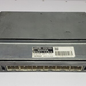 May include: A silver engine control module with a black border and a white label that reads "ENGINE CONTROL TOYOTA". The module has white connectors and mounting brackets.