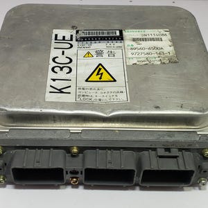 May include: A silver-colored electronic control unit (ECU) with a black and yellow warning label. The ECU has multiple gray connectors and the text "K13C-UE" is visible. The label includes Japanese text and a lightning bolt symbol.