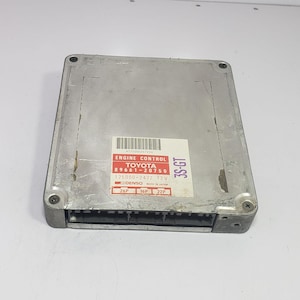 以下が含まれることがあります： バーコード、ENGINE CONTROL、TOYOTA 89661-20750、175000-2472 12V、DENSO、MADE IN JAPAN、3S-GT、およびピンラベル26P、16P、22Pのテキストが記載された白いラベルが付いた、長方形の銀色の金属製トヨタエンジンコントロールユニット。