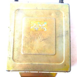 May include: A gold-coloured electronic control unit (ECU) with a small, yellow sticker and the Mitsubishi logo. The ECU has a bracket with two holes and two black plastic connectors. Likely for automotive use.