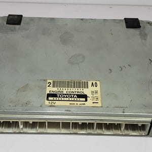 May include: A rectangular silver engine control module with a white label that reads "ENGINE CONTROL TOYOTA" and "MADE IN JAPAN". The module has white connectors on one side and two black rubber pads on top.