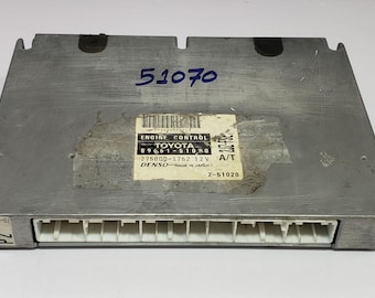 May include: A silver-colored engine control module with a rectangular shape. The module has a series of white connectors along one edge and a label with the text "ENGINE CONTROL TOYOTA" and other details. The number "51070" is written in blue.