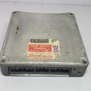 Può includere: Un modulo di controllo motore Toyota color argento con un connettore nero. Il modulo ha un adesivo con il testo "ENGINE CONTROL TOYOTA 89661-20712 175700-2832 12V" e altri dettagli.