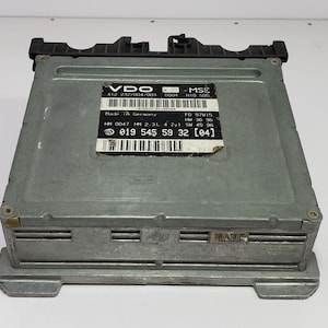 Puede incluir: Una unidad de control VDO MSE de color plateado con una parte superior de plástico negro. La unidad tiene una etiqueta con texto que incluye "Made in Germany" y un código de barras. La unidad es rectangular con ranuras horizontales.