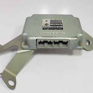 Puede incluir: Una unidad de control electrónico de color plateado con un soporte metálico. La unidad tiene forma rectangular con una serie de conectores y etiquetas. El soporte tiene orificios de montaje. El texto de la etiqueta incluye "NISSAN" y otros códigos.