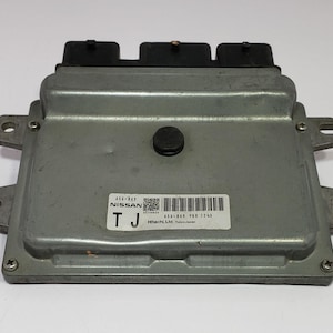 May include: A silver Nissan engine control unit (ECU) with black connectors. The ECU is rectangular with mounting tabs and a label showing the Nissan logo, a barcode, and the text "Hitachi Ltd., Tokyo Japan."