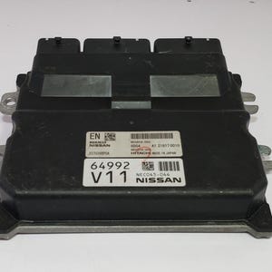 Puede incluir: Módulo de control del motor Nissan negro con múltiples conectores y soportes de montaje. El módulo tiene etiquetas con texto que incluye "EN", "NISSAN" y "64992 V11". Fabricado en Japón.