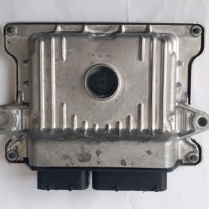 May include: A silver-colored electronic control unit (ECU) with a rectangular shape and a ridged top surface. It features two black connectors at the bottom and a circular black component in the center. The ECU is designed for automotive applications.