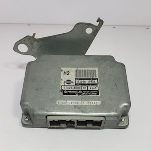 May include: A silver-colored electronic control unit with a bracket. The unit has a rectangular body with a connector and several labels with text and barcodes. The text includes "Nissan", "Hitachi, Ltd, Tokyo Japan", and model numbers.