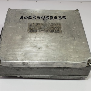 Puede incluir: Una unidad de control electrónico (ECU) de color plateado con el número A0235452235 escrito en la parte superior. Una pegatina con texto y un código de barras está adherida en la parte superior. La ECU es rectangular con esquinas redondeadas.