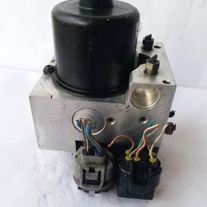 Op de afbeelding: Een zilveren en zwarte automotive ABS-module. De module heeft een zwart cilindrisch component aan de bovenkant en twee zwarte connectoren met gekleurde draden. De unit is ontworpen voor automotive gebruik.