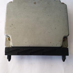 May include: A rectangular, silver-coloured electronic control unit (ECU) with a black plastic connector. The ECU has four gold screws and a few small spots on the surface. The black connector has two black plastic tabs.