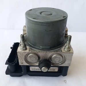 Op de afbeelding: Een grijze auto-ABS-module met een cilindrische bovenkant en een rechthoekige basis. De module heeft twee zilveren bouten en een zwarte connector aan de zijkant. De basis heeft een label met een streepjescode.