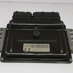 May include: Black Nissan engine control module with a rectangular shape and metal mounting brackets. The module has two black plastic connectors on top and a label with text and a barcode. The label reads "V 2".