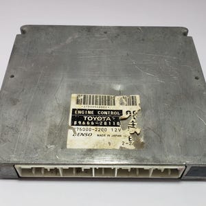 May include: A silver-colored engine control module with a rectangular shape. The module has a sticker with the text "ENGINE CONTROL TOYOTA" and other technical details. The module has a series of white connectors along one edge.
