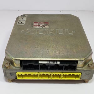May include: A silver ZEXEL electronic control unit (ECU) with black and yellow connectors. The ECU has the text "ZEXEL" printed on the top and a label with text and numbers. The unit has a rectangular shape.