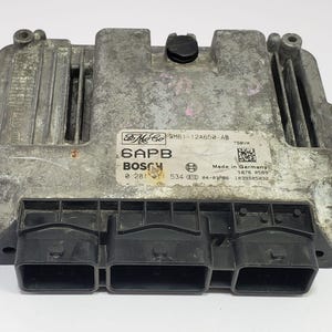 Puede incluir: Un módulo informático automotriz plateado con tres conectores negros. El módulo tiene una etiqueta con el texto "Ford", "Bosch" y "Made in Germany".