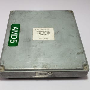 Puede incluir: Una unidad de control electrónico (ECU) rectangular de color plateado con una pegatina verde "AMD5". Una etiqueta blanca muestra "CONTROLLER" y otros códigos. La ECU tiene un código de barras y es probablemente para uso automotriz.