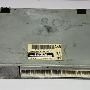 Puede incluir: Una unidad de control del motor de metal gris con una etiqueta negra que dice "ENGINE CONTROL TOYOTA 89661-37440 12V MADE IN JAPAN" y una etiqueta blanca que dice "C45 22411W05 J11-6".