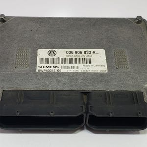 Puede incluir: Módulo informático automotriz rectangular plateado con dos conectores negros. El módulo tiene una etiqueta con el logotipo de Volkswagen, el texto "SIEMENS" y otra información técnica. Fabricado en Alemania.