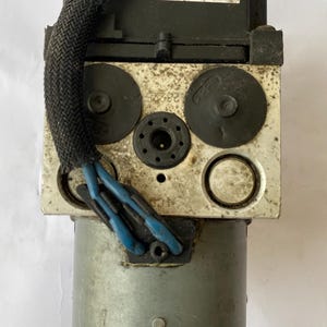 Op de afbeelding: Een auto-ABS-module met een zwarte bovenkant en een zilverkleurige behuizing. De module heeft meerdere poorten, connectoren en een label met het nummer 703511830872 060. Blauwe draden en een zwarte gevlochten kabel zijn bevestigd.
