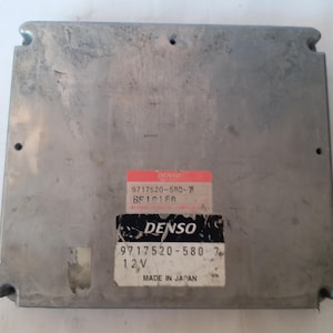 May include: A silver-coloured electronic control unit (ECU) with two labels. The top label is red and white, and the bottom label is white with black text. The text "DENSO" is visible on both labels, along with other numbers and the text "MADE IN JAPAN".