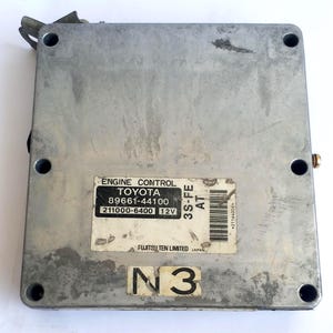 May include: A silver-colored engine control module with the text "ENGINE CONTROL TOYOTA" and model numbers. The module has several screw holes and a barcode. The text "FUJITSU TEN LIMITED JAPAN" is also visible.