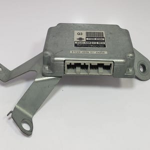 Könnte beinhalten: Ein silberfarbenes elektronisches Steuergerät mit angebrachtem Halter. Das Gerät hat eine rechteckige Form mit einer Reihe von Anschlüssen auf einer Seite und einem Etikett mit Text und einem Barcode. Die Halterung hat mehrere Befestigungslöcher.