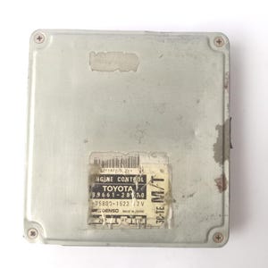 Könnte beinhalten: Ein rechteckiges, silberfarbenes Motorsteuergerät mit einem Etikett mit der Aufschrift "ENGINE CONTROL TOYOTA". Das Etikett enthält auch die Nummern "89661-2870" und "75802-1522 12V". Das Gerät ist mit vier Schrauben befestigt.
