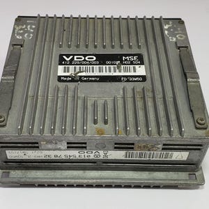 May include: A silver VDO MSE electronic control unit with a ribbed top and multiple labels. The labels include the text "Made in Germany" and various codes and numbers. The unit has a rectangular shape with metal clasps.