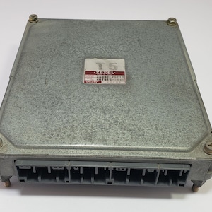 Op de afbeelding: Een zilverkleurige elektronische regeleenheid (ECU) met een rechthoekige vorm. De ECU heeft een label met de tekst "T5" en "ZEXEL". Het heeft meerdere connectoren en is vastgezet met vier schroeven. De unit is ontworpen voor automobieltoepassingen.