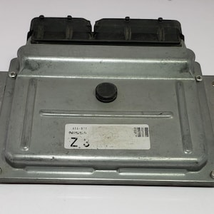 Puede incluir: Módulo de computadora automotriz plateado con conectores negros y una etiqueta "NISSAN Z3". El módulo es rectangular con pestañas de montaje y un botón negro central. Mide aproximadamente 20 cm x 15 cm.