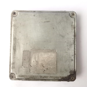 May include: A rectangular, silver-coloured electronic control unit (ECU) with four screws in the corners. The unit has a worn appearance with two rectangular stickers affixed to the surface. The ECU is likely for automotive or industrial applications.
