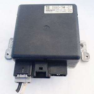 May include: A black electronic control module with multiple connectors and mounting brackets. The module has a label with text including "TB 39980-TFG -01" and other alphanumeric codes. Wires extend from one of the connectors.