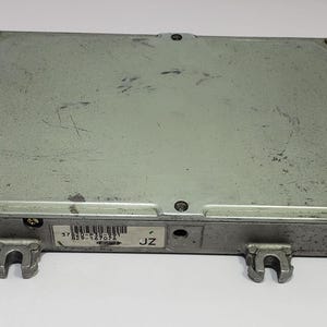 May include: A rectangular, silver-coloured electronic control unit (ECU) with a slightly textured surface. The ECU has a series of screws and mounting brackets. A sticker with text and a barcode is visible. Likely for automotive use.