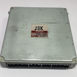 以下が含まれることがあります： 長方形の形状をした、銀色の電子制御ユニット（ECU）。ECUの片側には黒いコネクタがあり、「J8K」というテキストとその他の技術的な詳細が記載されたラベルが付いています。このユニットは自動車用途向けに設計されています。