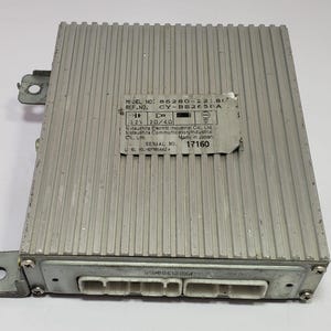 May include: A silver-colored electronic control unit with a ridged top surface. The unit has several connectors and mounting brackets. A label with text including "MODEL NO." and "SERIAL NO." is affixed to the top. The text "Made in Japan" is also visible.