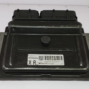 Może przedstawiać: Czarna elektroniczna jednostka sterująca (ECU) o prostokątnym kształcie z etykietą z napisem "NISSAN". ECU ma metalowe żebra z boku i mały, okrągły przycisk na górze. Urządzenie jest przeznaczone do zastosowań motoryzacyjnych.