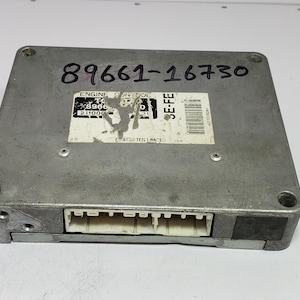 May include: A silver-colored engine control module with the numbers "89661-16730" written on top. The module has a white sticker with text and a barcode. It features a rectangular connector on one side.