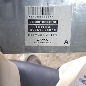 Könnte beinhalten: Ein silbernes Motorsteuergerät mit einem Etikett mit der Aufschrift "ENGINE CONTROL TOYOTA 89661-26B50". Das Etikett enthält auch den Text "DENSO MADE IN MALAYSIA".
