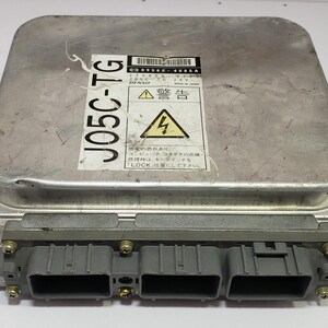 以下が含まれることがあります： 黒いコネクタと警告ラベルが付いたシルバー色の電子制御ユニット（ECU）。ラベルには日本語のテキストと稲妻のシンボルが含まれています。ユニットの側面には「J05C-TG」の文字が印刷されています。