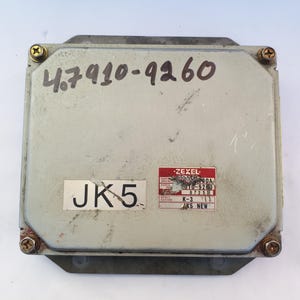 Può includere: Un'unità di controllo elettronica (ECU) rettangolare grigia, con segni di usura visibili. La superficie superiore presenta numeri scritti a mano e un adesivo con "JK5". È presente anche un adesivo rosso e bianco con il marchio "Zexel". Quattro viti fissano l'unità.