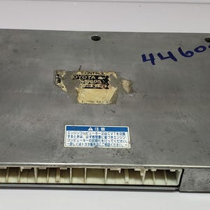 May include: A rectangular, silver Toyota engine control module with a row of white connectors. The module has a partially visible label and the number "44608" written in blue ink. A small blue and white label with Japanese text is also visible.