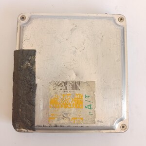 May include: A silver automotive computer module, rectangular in shape, showing signs of wear. A dark grey, textured component is partially detached on the side. A faded label with text and a barcode is attached to the front of the module.